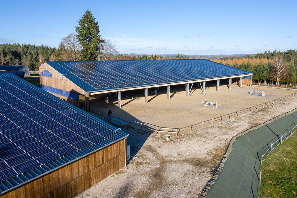 Bâtiment couvert avec panneaux solaires sur toit, entouré d'une piste en terre et d'une clôture, dans un environnement nat