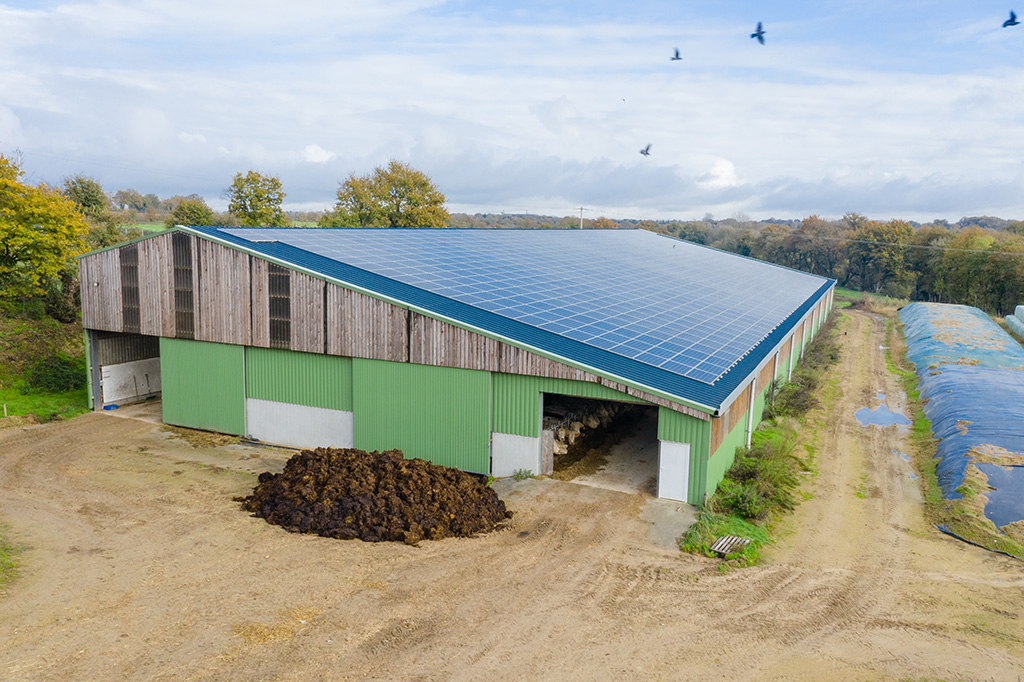 Bâtiment agricole avec toit solaire et compost à l'extérieur, entouré de champs et de végétation