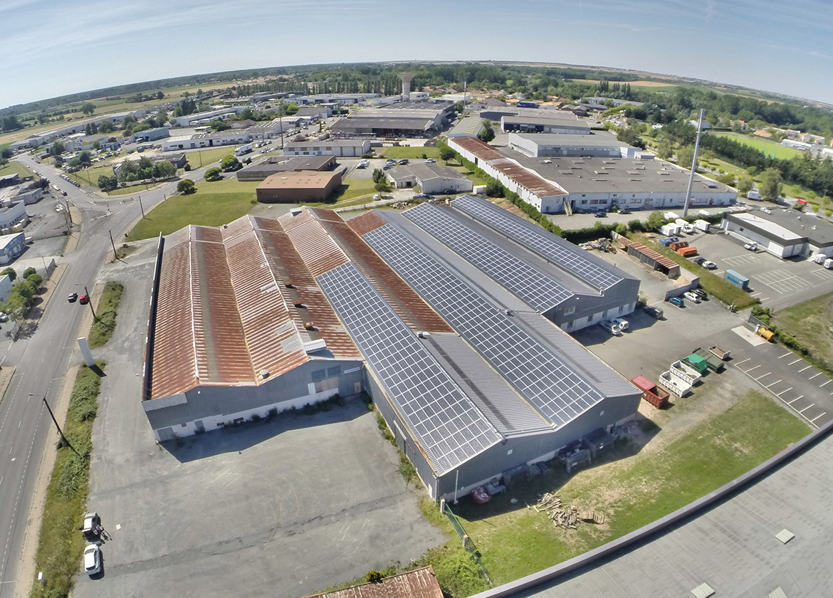 Vue aérienne d'un complexe industriel avec bâtiments couverts partiellement de panneaux solaires et parking adjacent