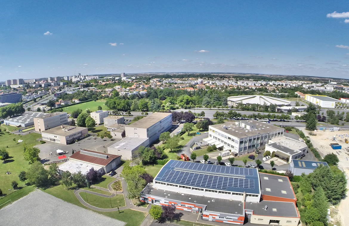 Vue aérienne d'un complexe industriel avec bâtiments et panneaux solaires, entouré de verdure et de quartiers résidentiel