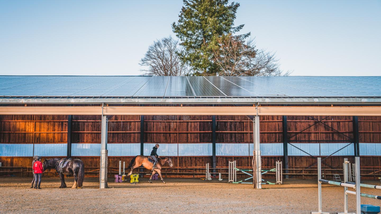 Centre équestre avec écuries modernes équipées de toiture photovoltaïque où un cavalier s’entraîne dans un manège e