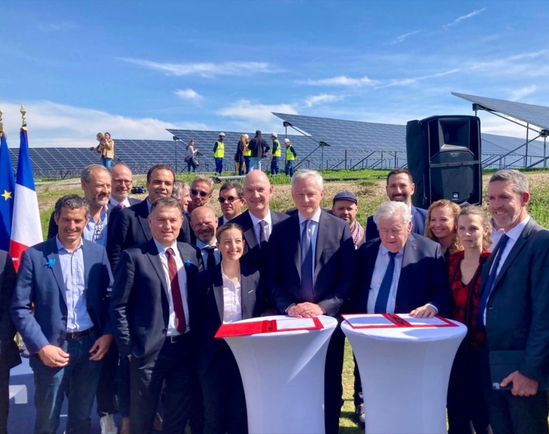 Groupe de personnes posant devant un champ de panneaux solaires lors d'une cérémonie officielle avec drapeaux français et