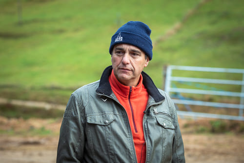 Homme portant un béret bleu et une veste grise sur un fond naturel avec une clôture en arrière-plan