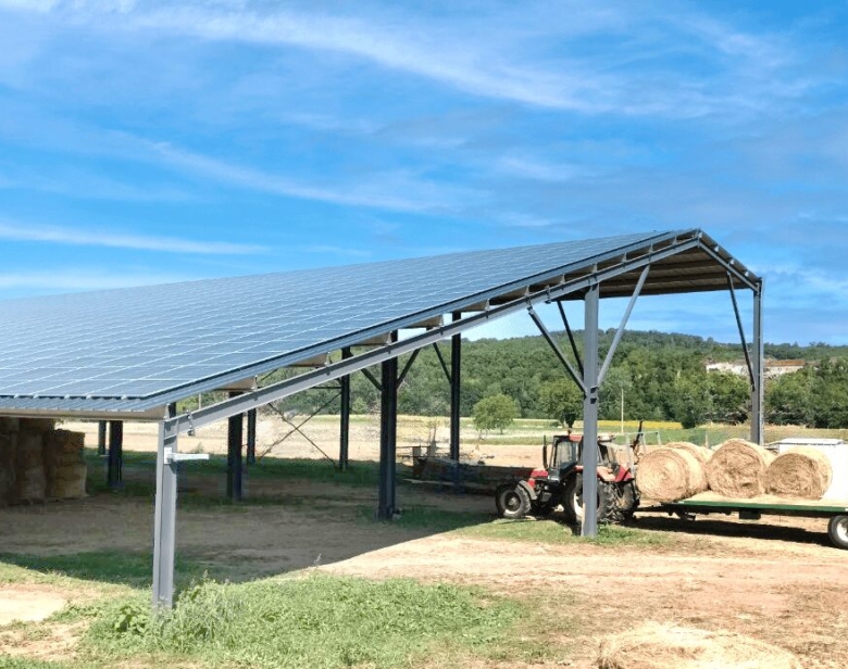Abri métallique avec panneaux solaires abritant un tracteur et une remorque chargée de balles de foin dans un champ
