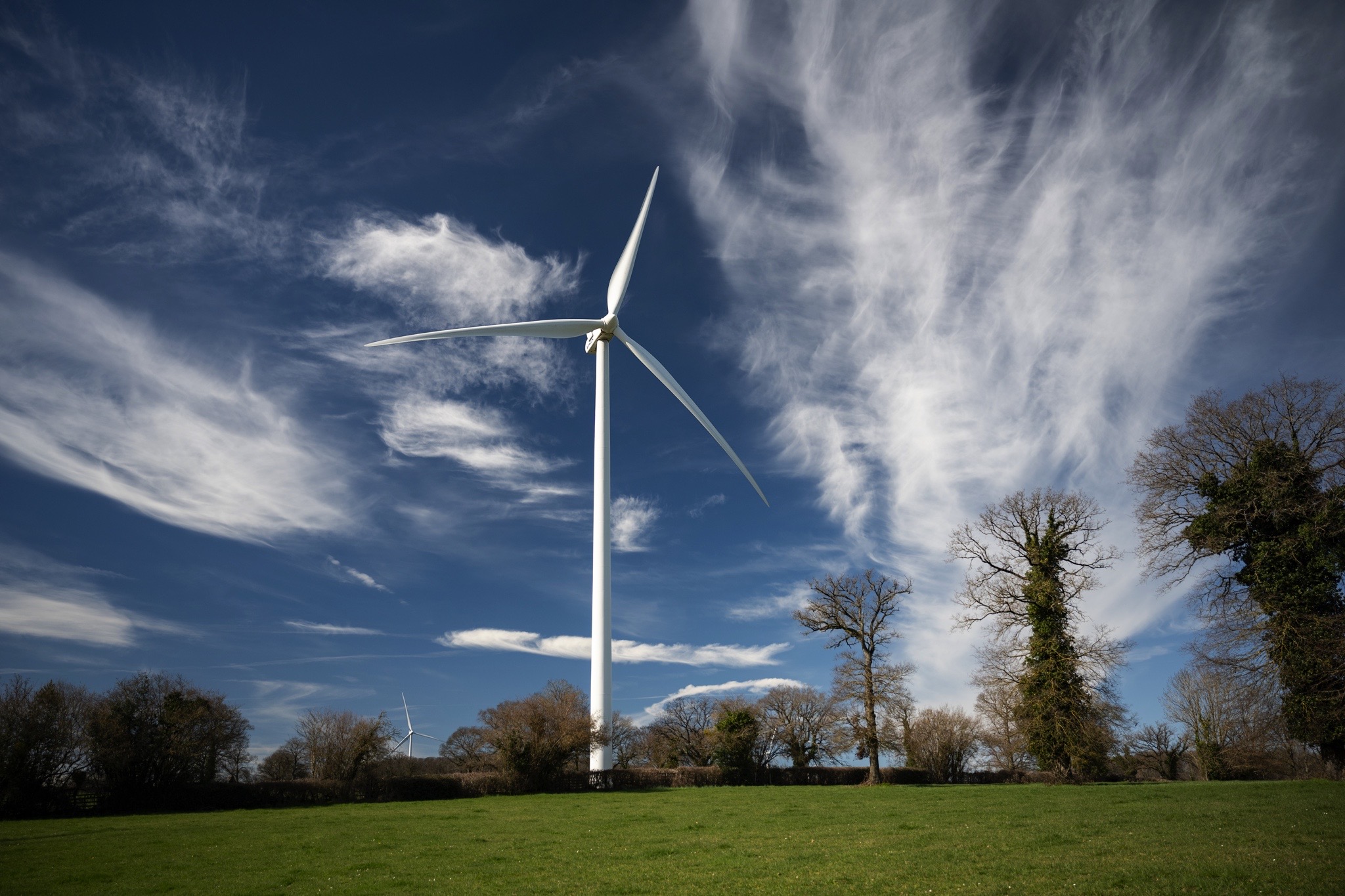 Éolienne moderne dans un champ vert sous un ciel bleu avec nuages dispersés et arbres en arrière-plan
