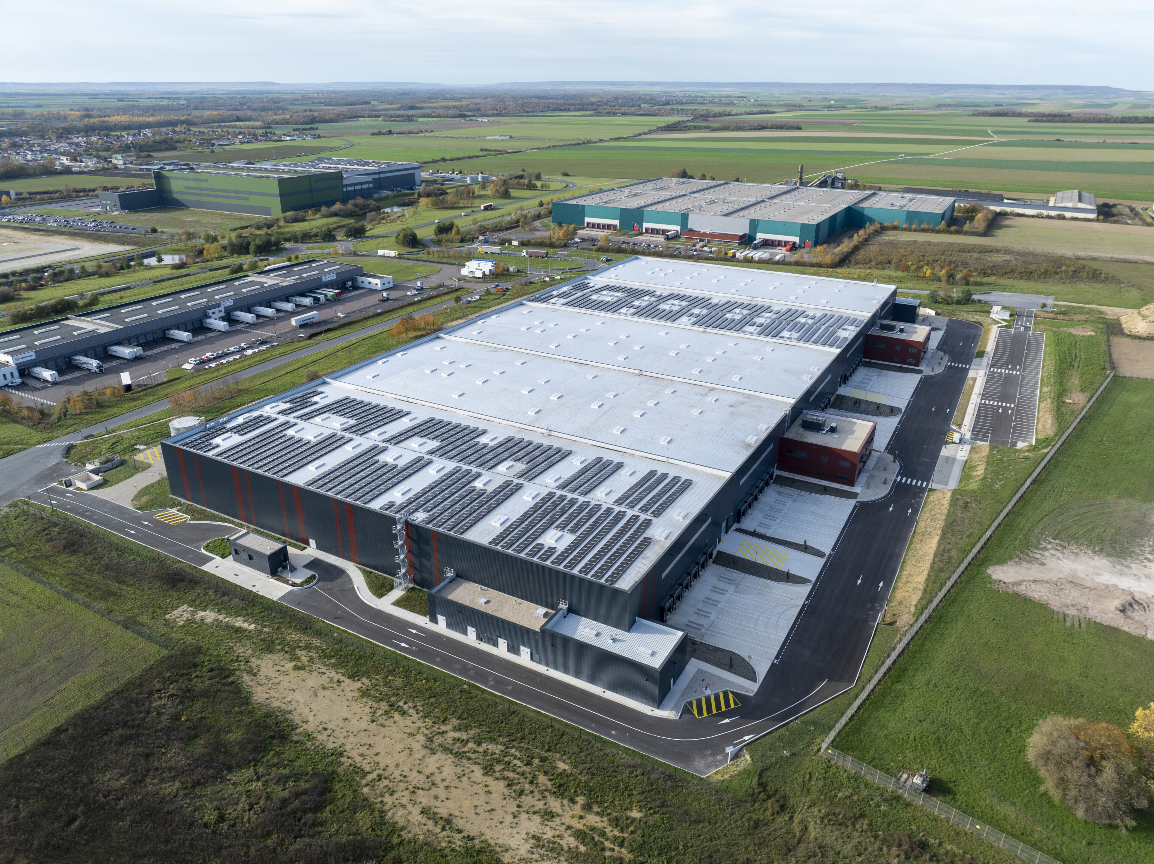 Vue aérienne d’un vaste complexe industriel avec des bâtiments de stockage et des parkings adjacents en zone rurale entou